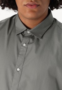 Camisa gris de botones con una textura suave, que presenta un cuello y botones blancos a lo largo de la tapeta. Vista de cerca que se centra en la parte superior del torso.