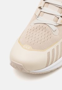 Beige idrottssko med en texturerad mesh-överdel, släta syntetiska överlägg och vit sula. "EA7"-logotyp synlig nära tån.