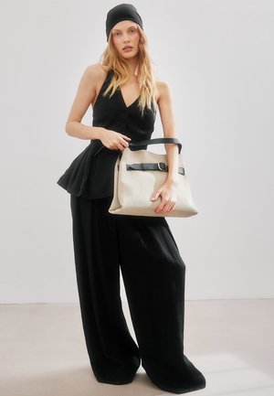 Mlada ženska v črni majici brez rokavov, širokih črnih hlačah in beanieju drži veliko bež torbo s črnim trakom na svetlem ozadju.