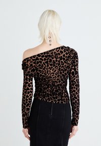 Barna leopárdmintás felső hosszú ujjal, vállra eső kialakítással és ráncolt részletekkel. A textúra puhanak és enyhén fényesnek tűnik. Fekete szoknya látható alatta.