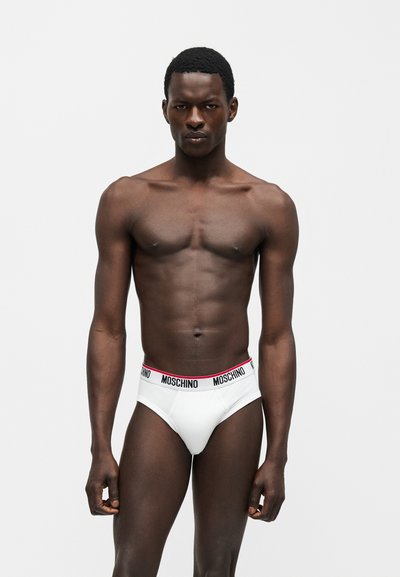 Άνδρας μοντέλο με σκοτεινό δέρμα που φοράει λευκά εσώρουχα Moschino με μαύρη και κόκκινη ζώνη στη μέση, στέκεται μπροστά σε απλό φωτεινό φόντο.