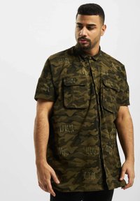 Camicia a maniche corte, con bottoni, in camouflage verde con dettagli dorati e due tasche sul petto, caratterizzata da una vestibilità rilassata e tessuto testurizzato.