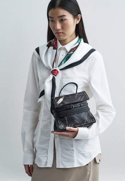 Woman wearing white shirt and colorful scarf holds black leather handbag with white stitch details and small feather charm.