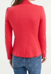 Korálový přiléhavý blejzr s dlouhými rukávy, strukturovanými rameny a hladkou texturou. Zadní část má centrální šev a mírně zakřivený lem.