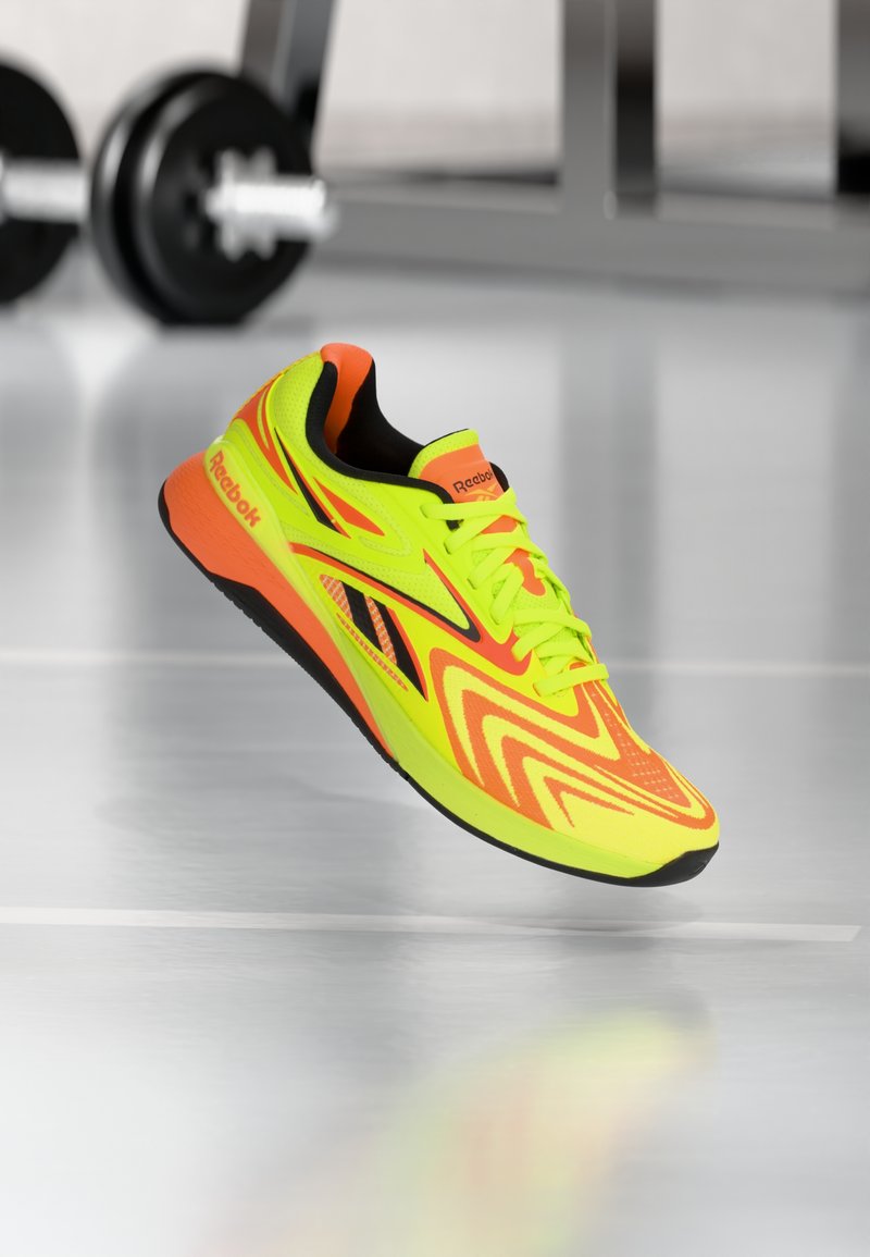 Ένα παπούτσι για τρέξιμο Reebok σε νέον κίτρινο και πορτοκαλί με μαύρη σόλα, αιωρείται πάνω από αντανακλαστικό πάτωμα γυμναστηρίου, με θολά βάρη στο φόντο.