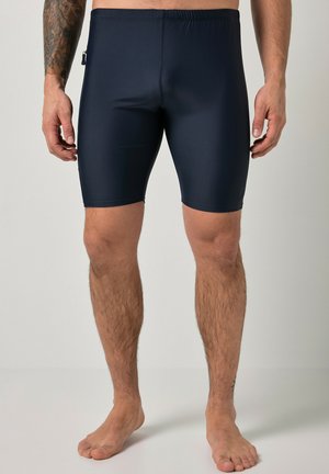 Mann trägt schwarze enge knielange Badeshorts und steht barfuß vor einem einfarbigen Hintergrund.