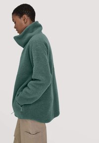 Green fleece pullover with a high collar, side zip, and front pocket. Soft texture, relaxed fit, and curved hem design.