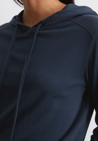 Marineblauwe hoodie met een zachte textuur, voorzien van een trekkoord in de capuchon en een relaxte pasvorm, die de minimalistische stijl en strakke lijnen benadrukt.