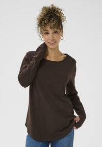 Brun langærmet skjorte lavet af blødt stof, med rund halsudskæring, slim fit og let buet kant. Bæres med afslappede blå jeans.