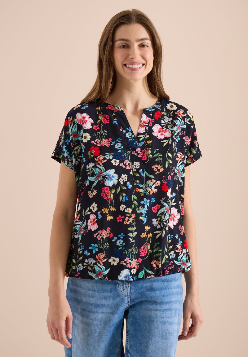 Jeune femme souriante, portant un chemisier noir à manches courtes à fleurs et un jean bleu clair sur un fond beige.
