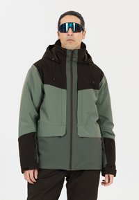 Wasserabweisende Jacke in Grün und Schwarz, mit Reißverschluss, verstellbarem Kapuzenbund, zwei Fronttaschen und strukturiertem Stoff. Große Sonnenbrille getragen.