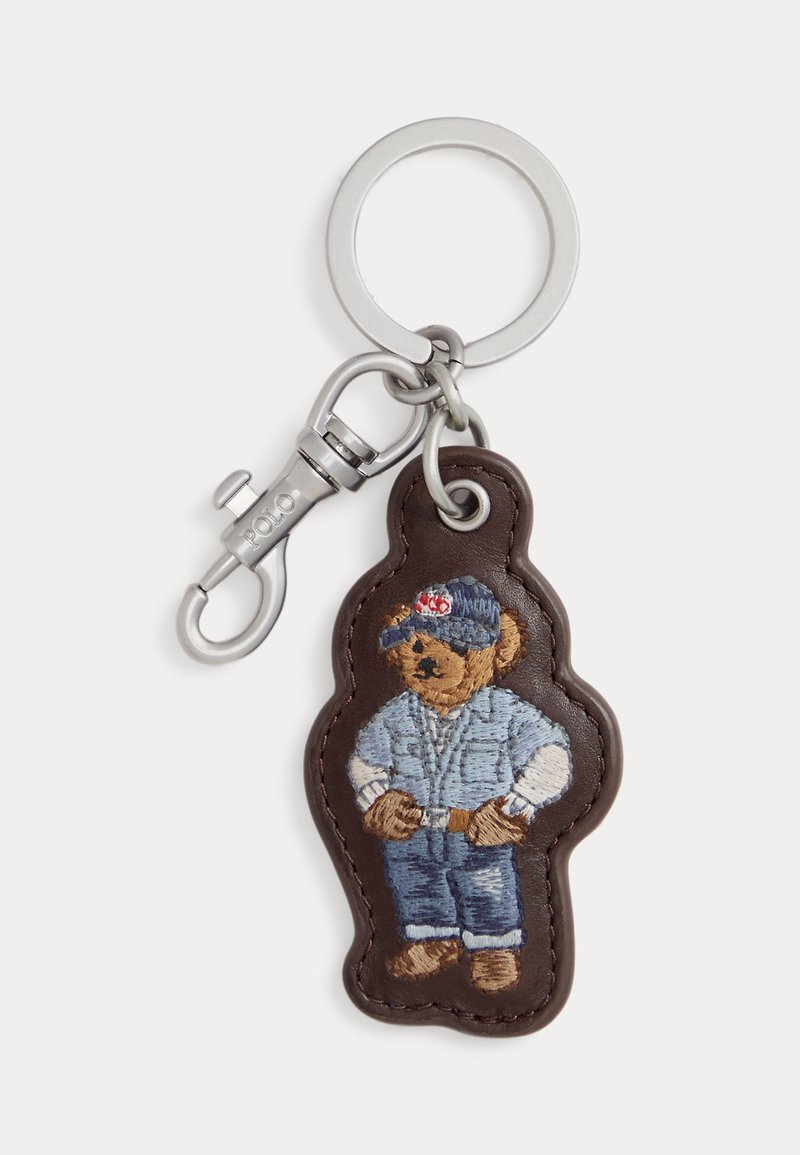 Porte-clés en métal avec une étiquette en cuir marron brodée d'un ours portant une casquette bleue, une veste, un jean et des chaussures marron.