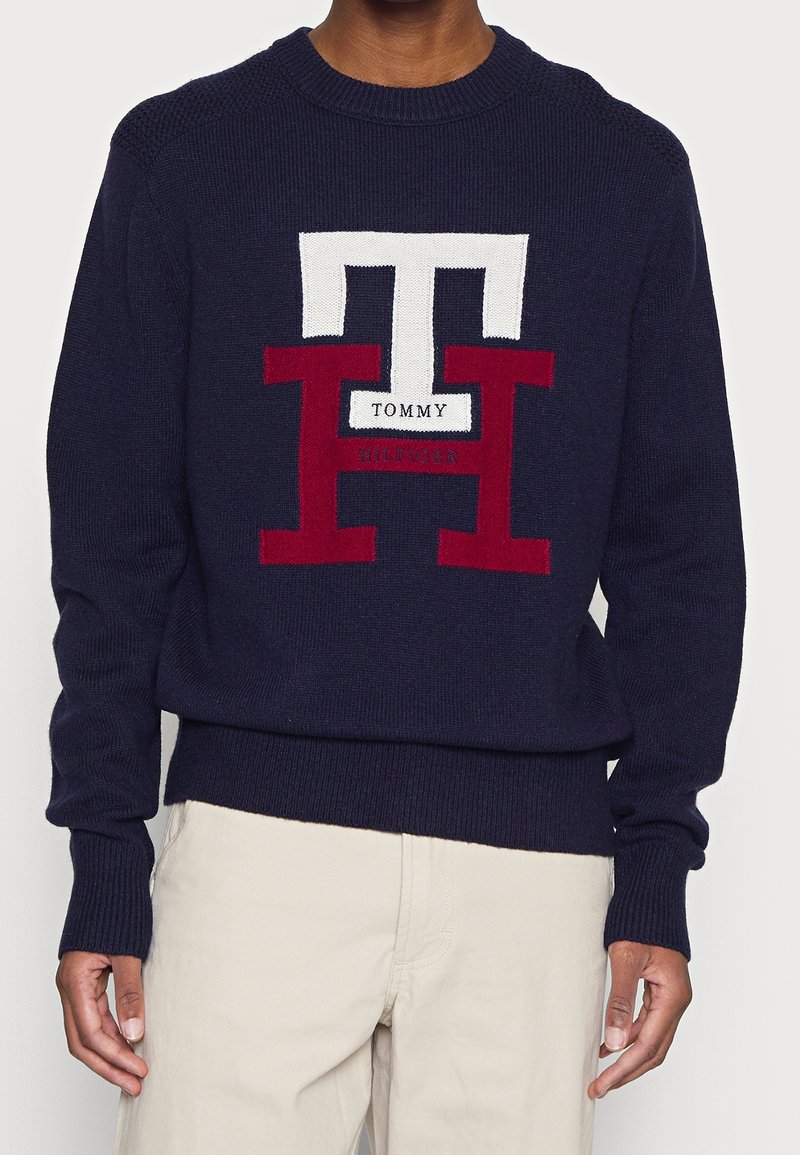 Personne portant un pull en maille bleu marine avec de grandes lettres blanches et rouges du logo Tommy Hilfiger, associé à un pantalon beige clair.