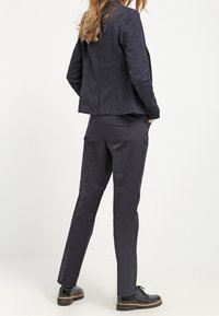 Marinblå prickig blazer med en figursydd design som är kombinerad med mörka byxor. Skorna är blanka svarta med en texturerad sula. Enkel, modern siluett.