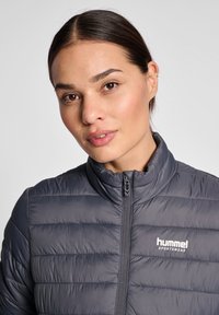 Grå pufferjacka med horisontellt sydd mönster, hög krage och framdragkedja. Har "hummel"-logotypen i vitt på vänster bröst.
