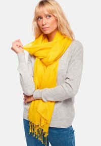 Femme blonde portant un pull gris clair et un jean bleu tient une écharpe jaune vif enroulée autour de son cou sur un fond blanc.