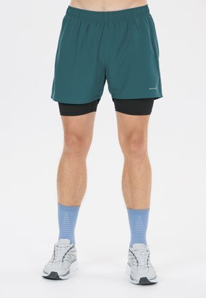 Man draagt teal hardloopshorts met zwarte compressieshorts eronder, lichtblauwe sokken en witte hardloopschoenen op een witte achtergrond.