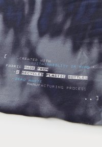 Recyklovaná tkanina označená tmavým tie-dye vzorom, s bielym a modrým textom, ktorý vysvetľuje udržateľný zdroj materiálov a nulový odpad.