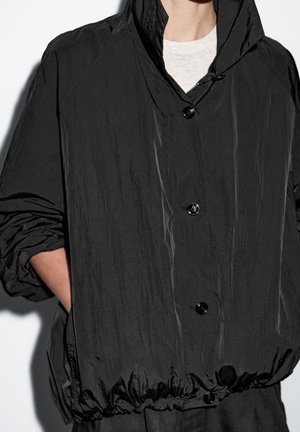 Veste noire à capuche avec boutons à l’avant et taille élastique, portée sur une chemise claire, les mains dans les poches latérales.
