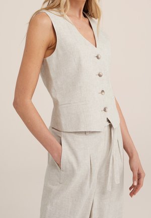 Kvinde iført ærmeløs beige hørvest med fire knapper og matchende høj taljebukser med lommer, mod en ensfarvet baggrund.