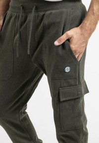 Pantalons cargo vert olive en tissu texturé, avec une taille élastique dotée de cordons de serrage et une poche cargo latérale avec rabat.
