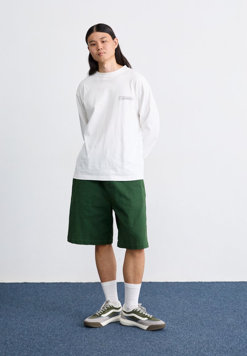 Witte langemouwen shirt met een klein logo, gecombineerd met groene shorts. Het schoeisel bestaat uit grijze en groene sneakers, aangevuld met witte sokken.