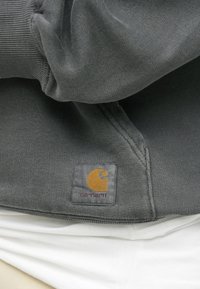 Felpa grigia con texture a coste; presenta un logo rettangolare ricamato in giallo e grigio, posizionato in basso a sinistra.