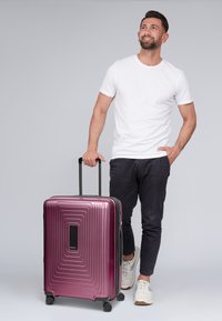 Valigia rigida di colore bordeaux lucido con linee orizzontali testurizzate, manico nero e ruote, esposta accanto a una persona in abbigliamento casual.