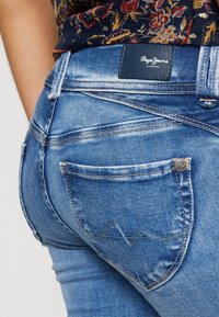 Blue denim jeans with a close-up view. Features include a dark leather brand tag, slight fading, and subtle stitching patterns on the pockets.
