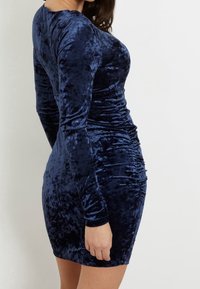 Mörkblå sammetklänning med långa ärmar och draperad detalj på sidan, med en figurnära passform och mjukt, strukturerat tyg.