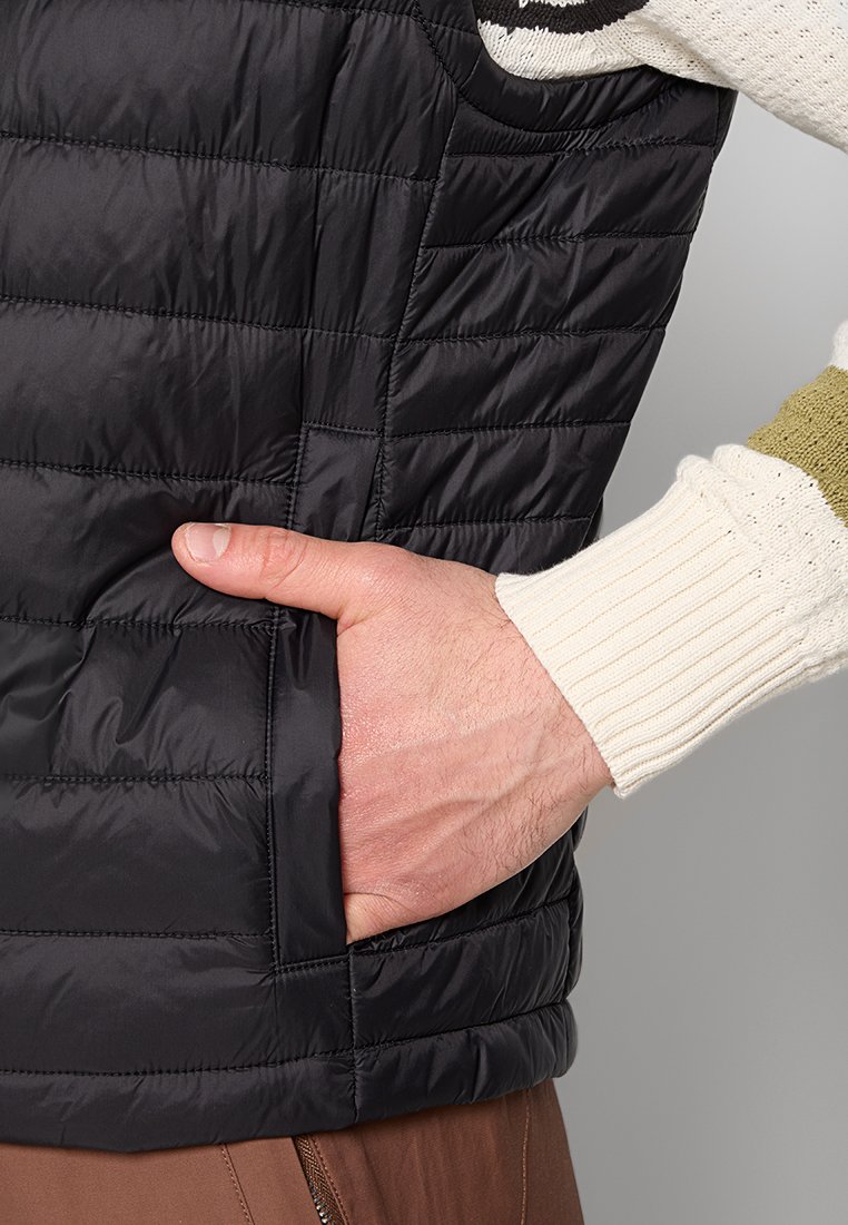 Gilet nero imbottito con motivo quilted, materiale leggero, tasca laterale, indossato sopra un maglione crema con polsini a coste.