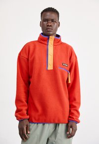 Rød fleecepullover med en orange knaplukning og blå detaljer. Har en lynlåslomme og tekstureret stof.