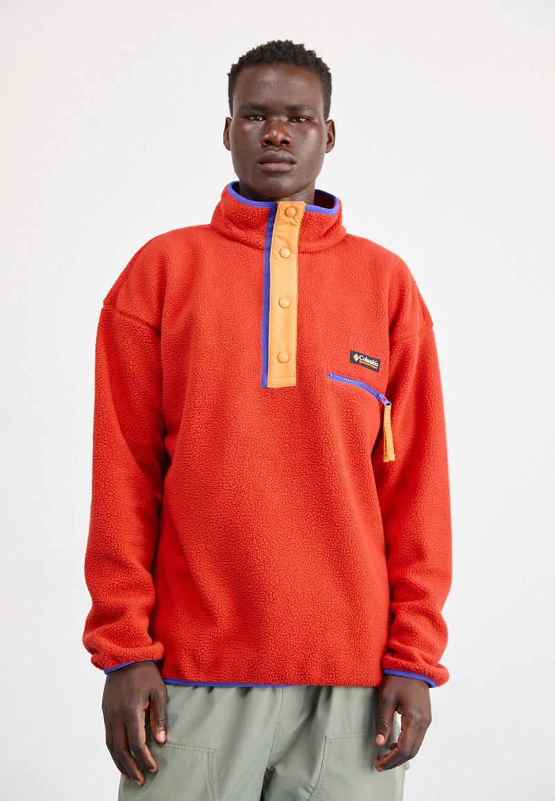 Rød fleecepullover med en orange knaplukning og blå detaljer. Har en lynlåslomme og tekstureret stof.