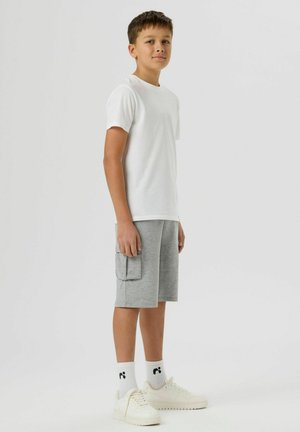 Jongen staand in zijaanzicht, gekleed in een wit T-shirt, grijze cargoshorts, witte sokken en witte sneakers, tegen een effen lichte achtergrond.