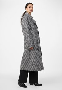 Cappotto lungo grigio e nero a spina di pesce con ampio risvolto e vita stretta, abbinato a pantaloni neri a gamba larga e stivaletti.