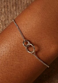 Zilveren kettingarmband met twee met elkaar verbonden ringen, tegen een achtergrond van textuur huid met een warme tint. Minimalistisch ontwerp en een gladde afwerking.