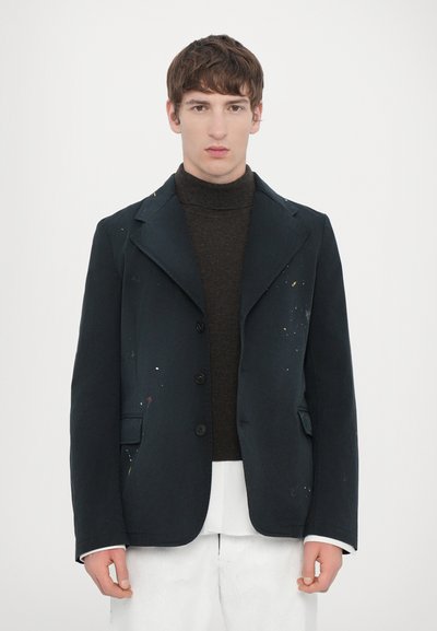 Chaqueta entallada de color azul marino, con un frente de tres botones, bolsillos laterales y detalles de salpicaduras de pintura, llevada sobre un suéter de cuello alto marrón.