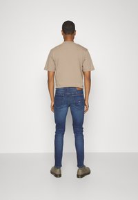 Béžové bavlněné tričko, modré slim-fit džíny a šedé boty. Viditelné zadní kapsy na džínech, jednoduchý design a hladká textura.