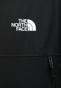 Zwarte zip-up jack met een wit logo van "THE NORTH FACE" op de bovenste linkerkant. Het materiaal lijkt soepel met een matte textuur.