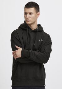 Hoodie en coton noir avec une poche kangourou à l'avant, des cordons de serrage ajustables et un logo blanc "A.R.C." sur la poitrine gauche. Texture douce.