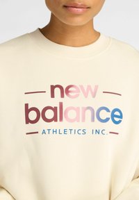 Sweatshirt couleur crème avec un imprimé graphique "new balance" en dégradé de rose et de bleu, et "ATHLETICS INC." en texte bleu en gras.