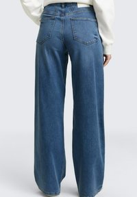 Persoon die wijd uitlopende blauwe spijkerbroek en een wit shirt met lange mouwen draagt, gezien van achteren tegen een effen achtergrond.