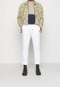 Geruite groene en beige shirt, lichtgrijs T-shirt, witte broek met rode accenten, zwarte veterschoenen en een kleine zwarte pouch tas.