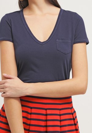 Frau trägt ein marineblaues T-Shirt mit V-Ausschnitt und Brusttasche sowie einen roten Rock mit marineblauen horizontalen Streifen, die Arme vor dem Körper verschränkt.