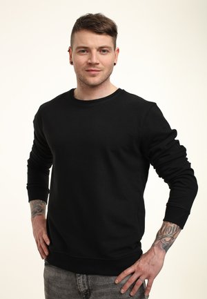 Jongeman met kort haar, zwarte oorbellen, een zwart sweatshirt en getatoeëerde onderarmen staat met handen op de heupen tegen een witte achtergrond.
