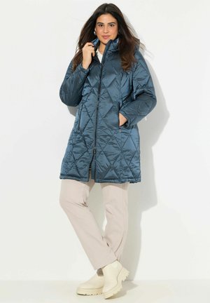 Quilted, mørk teal pufferjakke med hætte og frontlynlås, der har sidelommer og en knælang skæring, parret med beige bukser og ankelstøvler.