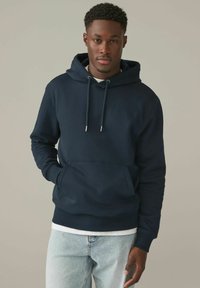 Sudadera azul marino con un bolsillo frontal, capucha con cordón y puños de canalé. Llevada sobre una camiseta blanca y combinada con jeans de lavado claro.