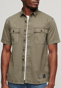 Camisa de manga corta con cuello en algodón verde oliva. Cuenta con dos bolsillos en el pecho, cierre de botones en la parte delantera y una etiqueta en el lado inferior izquierdo.