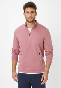 Uomo che indossa un pullover rosa polveroso con mezza zip e tasche laterali con zip, pantaloni blu navy, in piedi contro uno sfondo bianco semplice.