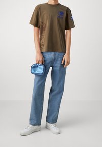 Camiseta de algodón verde oliva con un gráfico del globo y texto azul, combinada con unos jeans de mezclilla azul claro y zapatillas blancas, sosteniendo un bolso azul brillante.